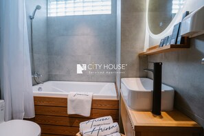 A bathroom with a bathtub, a shower, a toilet, and a sink. There is a wooden vanity with a towel and a sign that says "CityHouse Managed by CityHouse". - CityHouse VIBE Abora (Ho Chi Minh City)