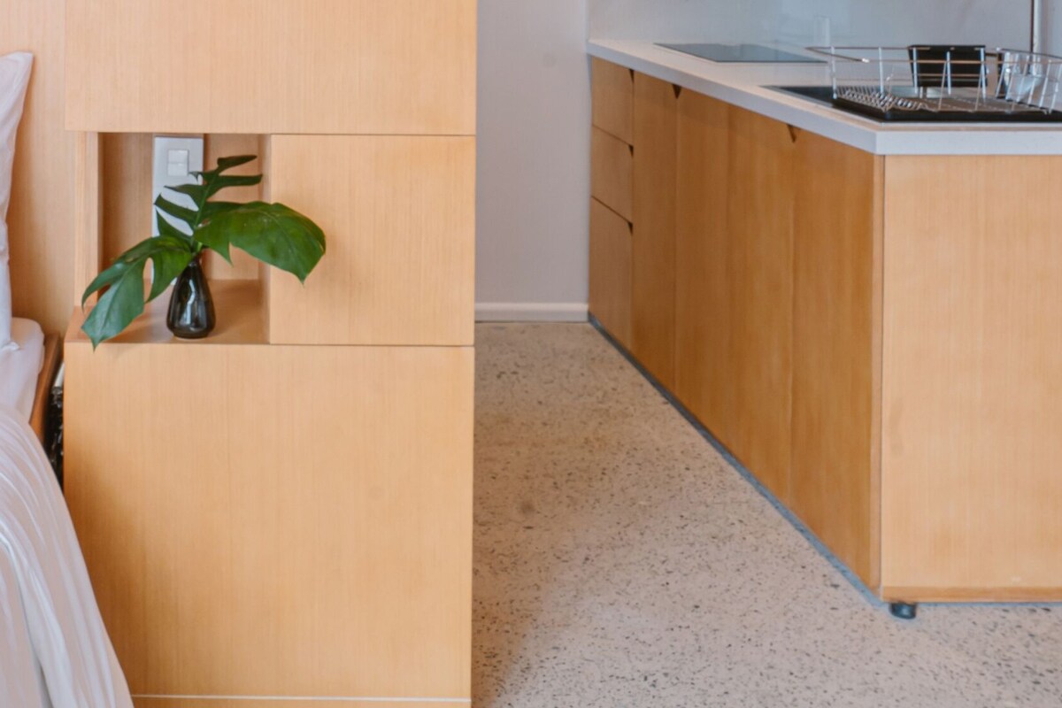 A bedroom with a bed, a nightstand, a small cabinet with a plant, and a kitchenette area.