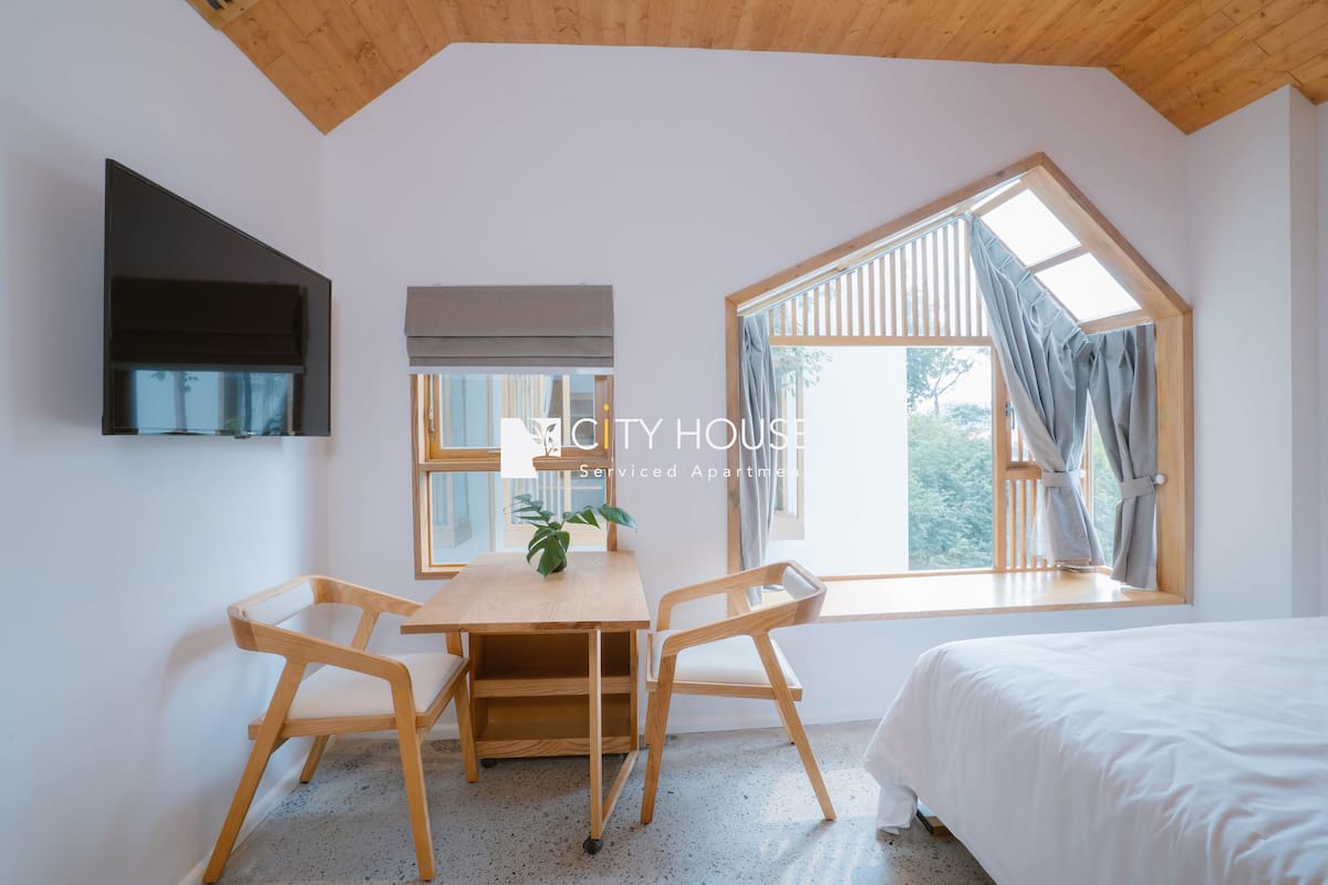 Un dormitorio con cama, escritorio, dos sillas, una planta en maceta y ventanas amplias.