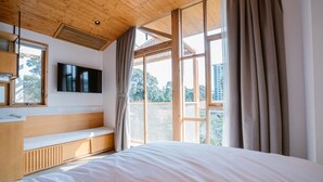 Un dormitorio con techo de madera, un ventanal con cortinas, televisor montado en la pared y cama con colchón blanco.