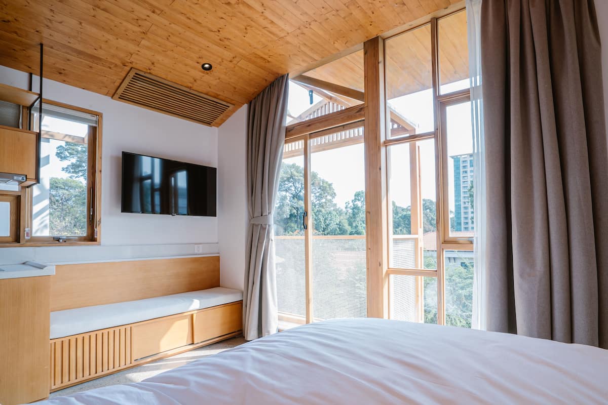 Un dormitorio con techo de madera, un ventanal con cortinas, televisor montado en la pared y cama con colchón blanco.