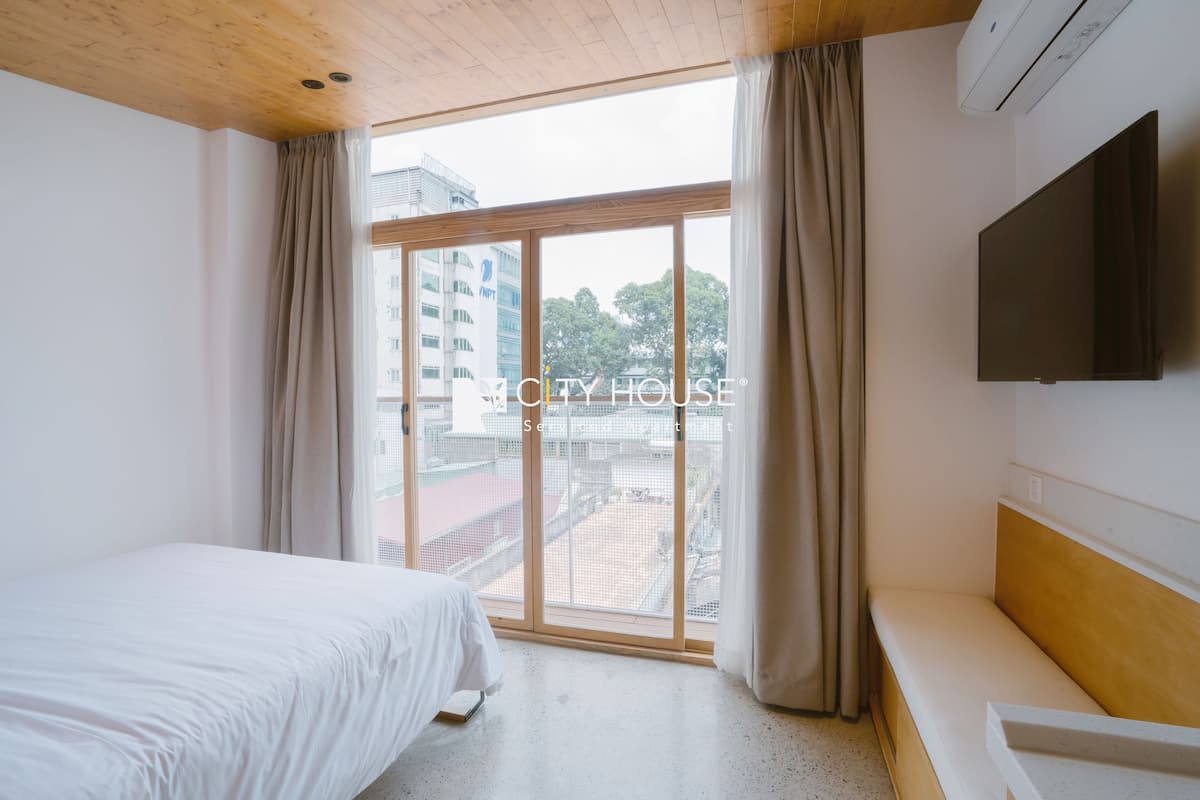 Habitación de hotel con cama, un banco, televisor y un amplio ventanal con cortinas.