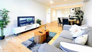 A modern living room with a flat-screen TV, a wooden coffee table, a grey sofa, and a dining area in the background.
