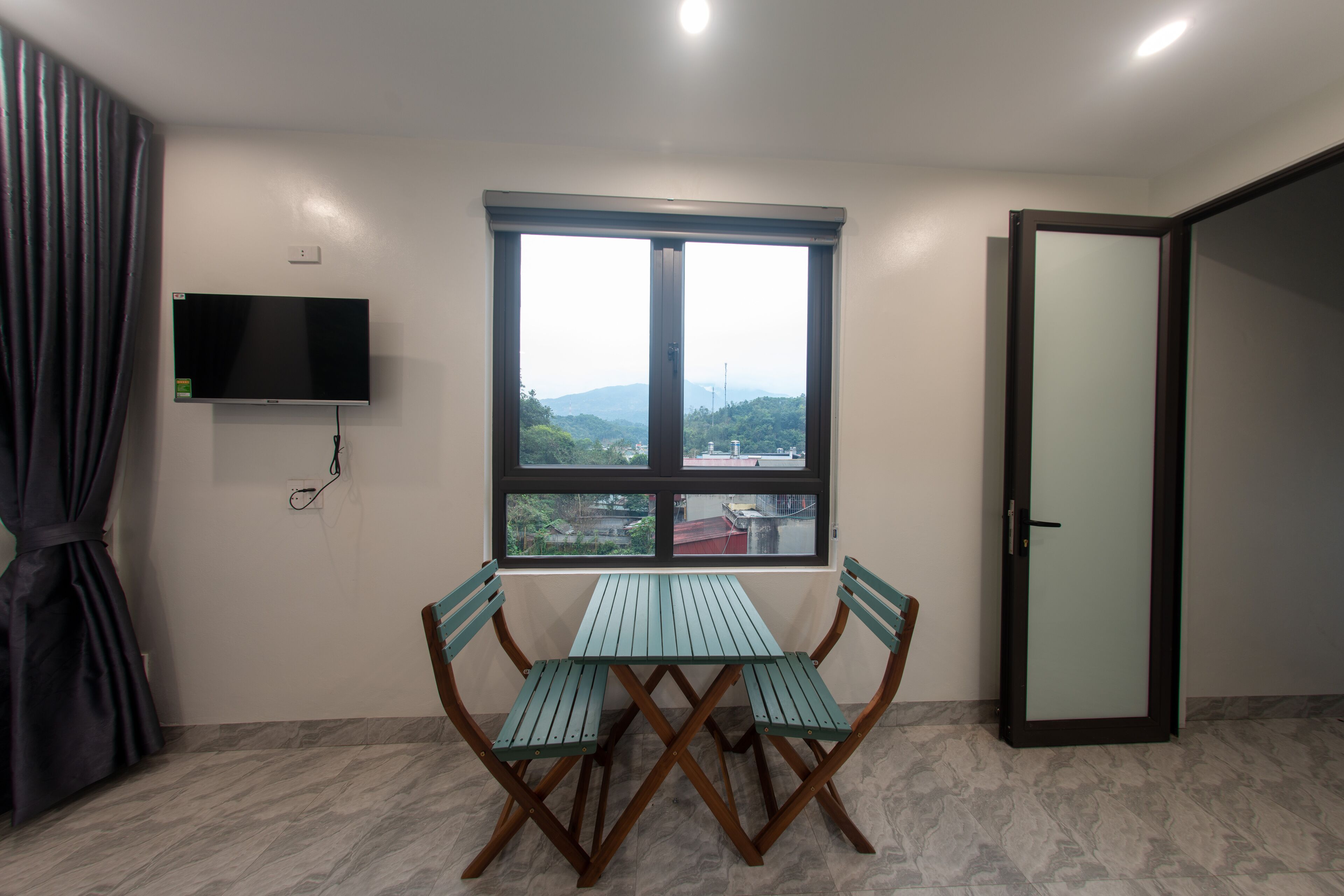 A room with a table and chairs, a TV mounted on the wall, and a window with a view of greenery and buildings.