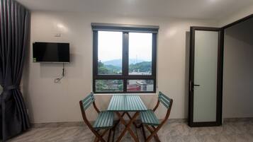 A room with a table and chairs, a TV mounted on the wall, and a window with a view of greenery and buildings.
