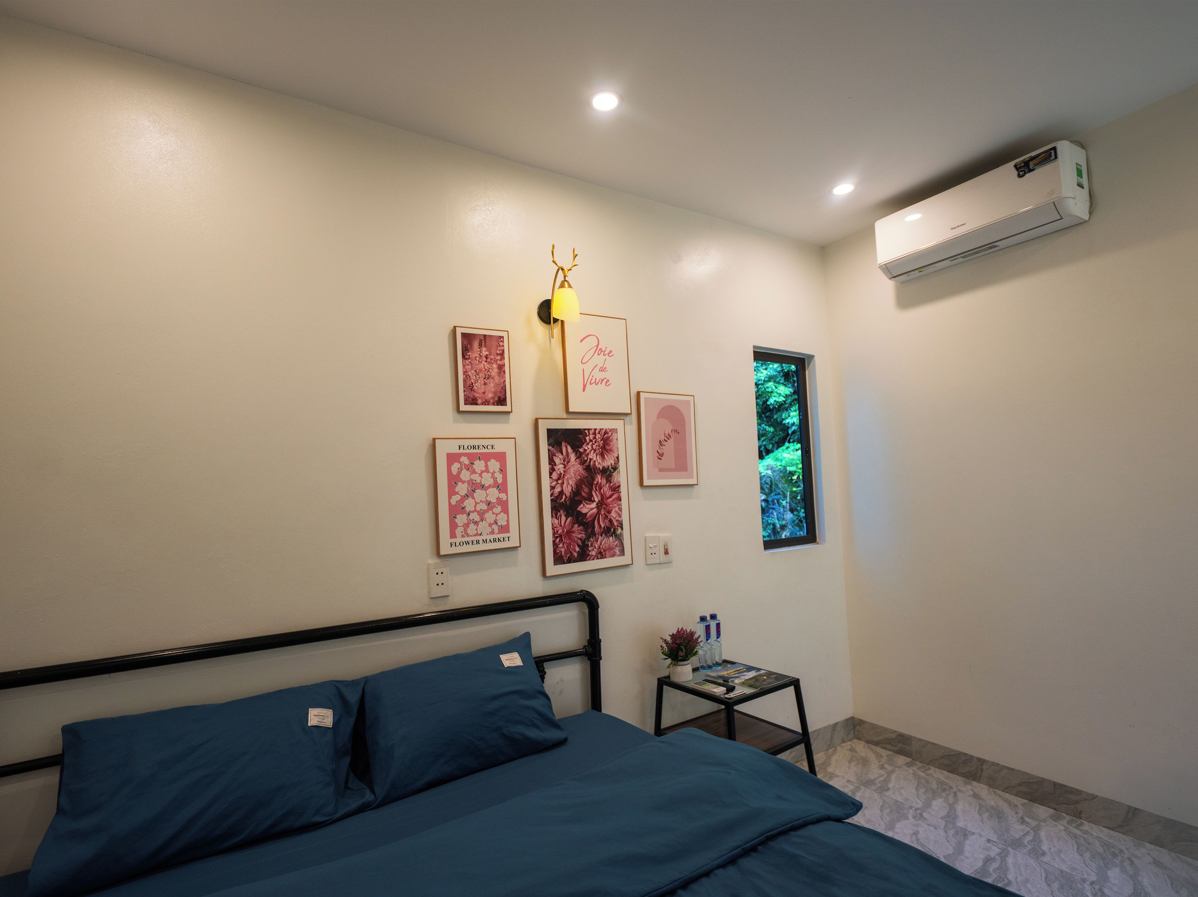 A bedroom with a bed, a small table with a plant and water bottles, a wall-mounted air conditioner, and framed pictures on the wall.