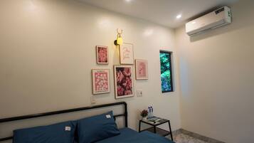 A bedroom with a bed, a small table with a plant and water bottles, a wall-mounted air conditioner, and framed pictures on the wall.