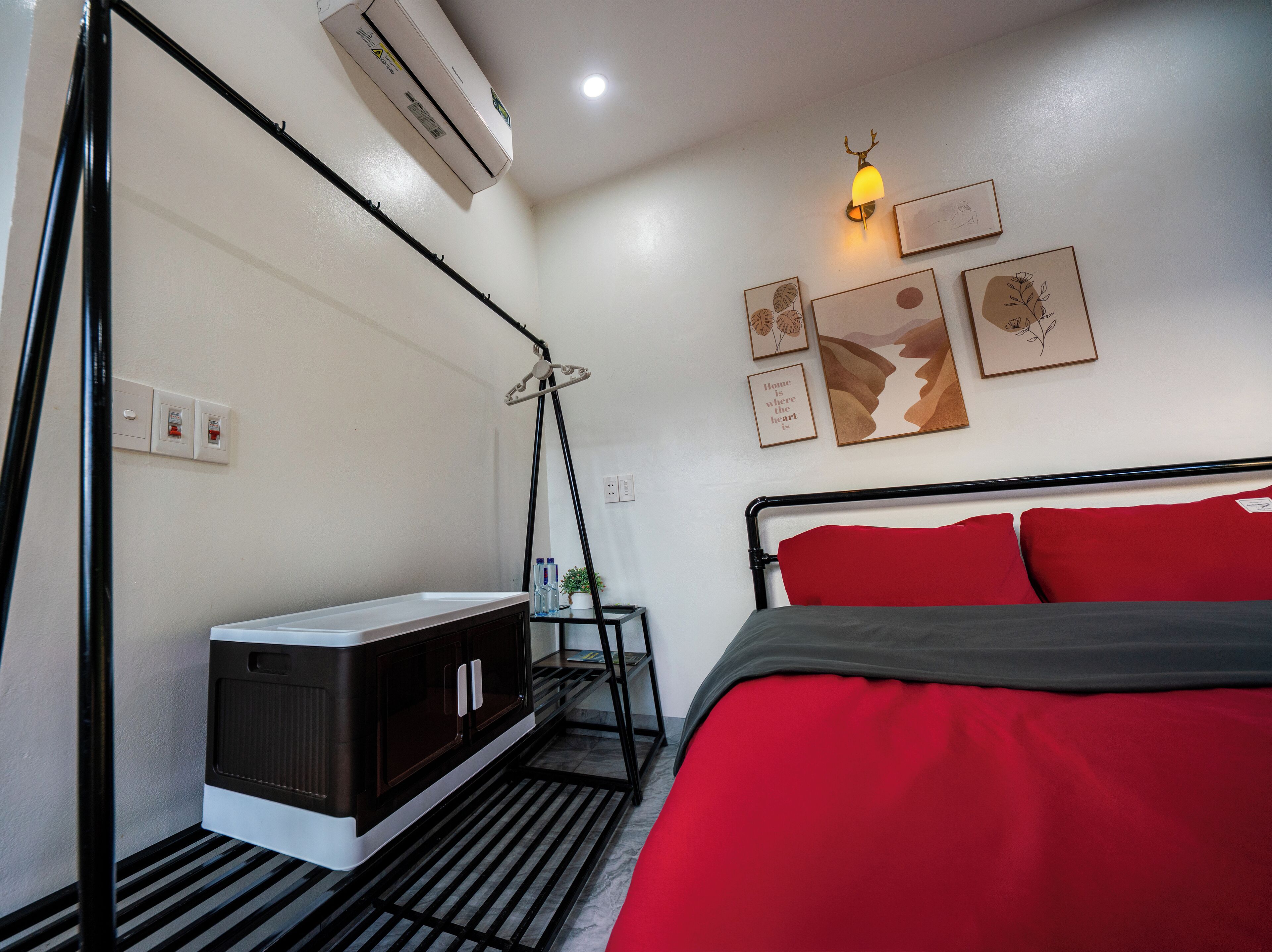 A bedroom with a bed, a bedside table, an air conditioner, a wall-mounted lamp, and framed artwork.