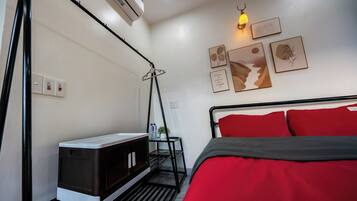 A bedroom with a bed, a bedside table, an air conditioner, a wall-mounted lamp, and framed artwork.