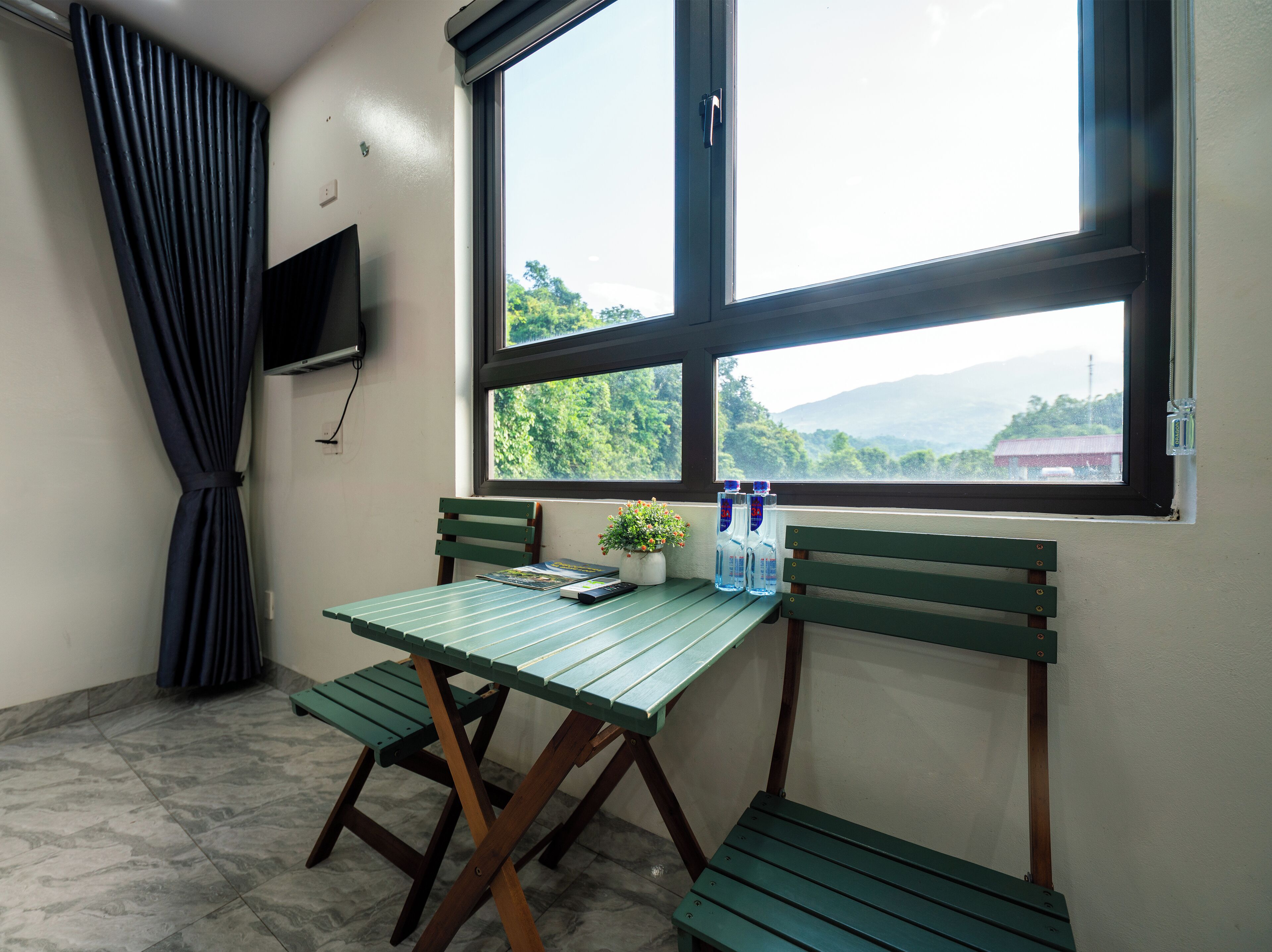Una habitación con una mesa y banco verdes, una planta en maceta y una ventana con vistas a vegetación y montañas.