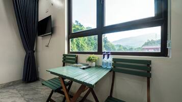 Una habitación con una mesa y banco verdes, una planta en maceta y una ventana con vistas a vegetación y montañas.