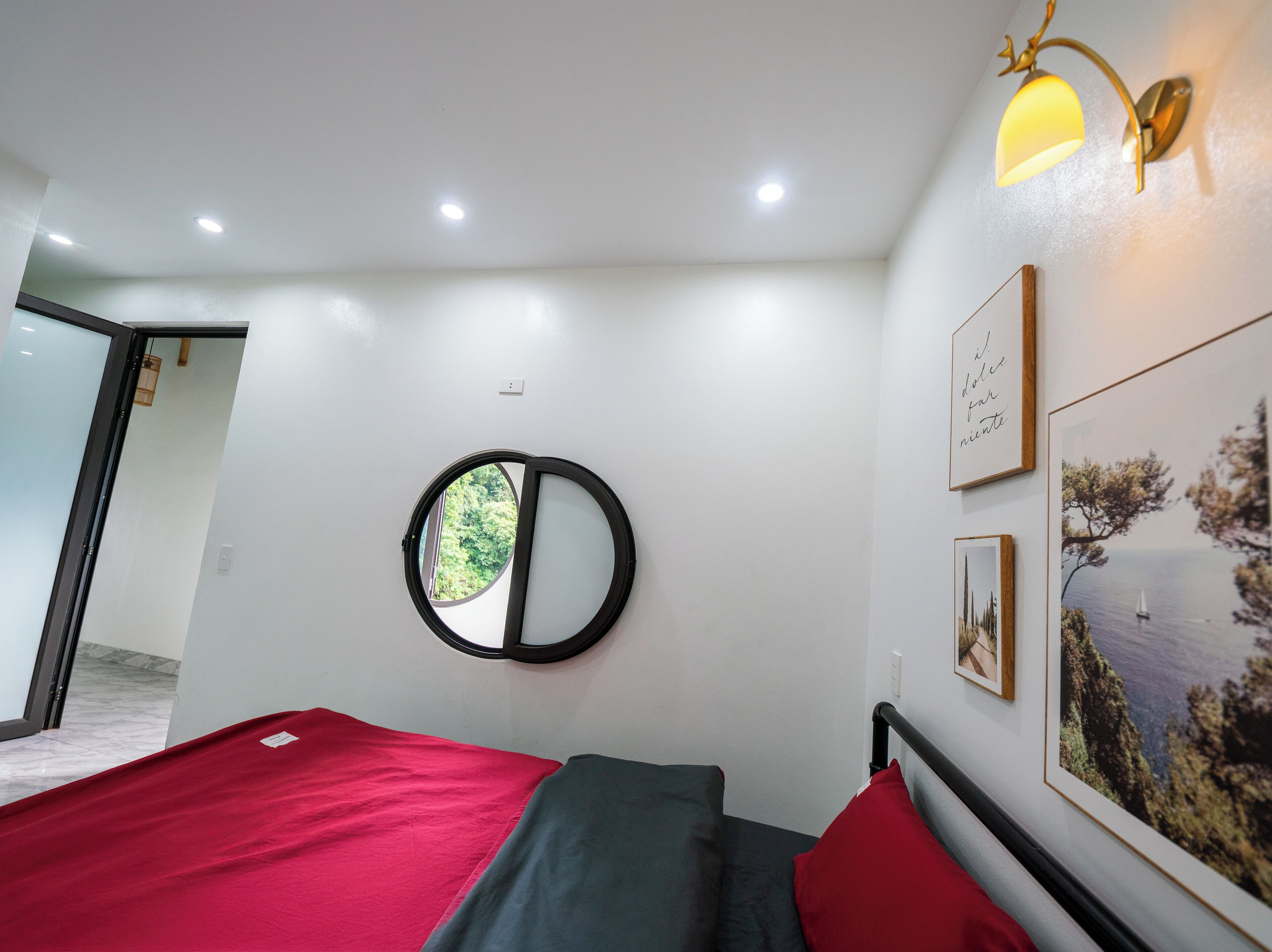A room with a bed, a round mirror, and a window with a view of greenery.