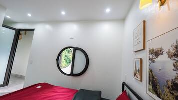 A room with a bed, a round mirror, and a window with a view of greenery.