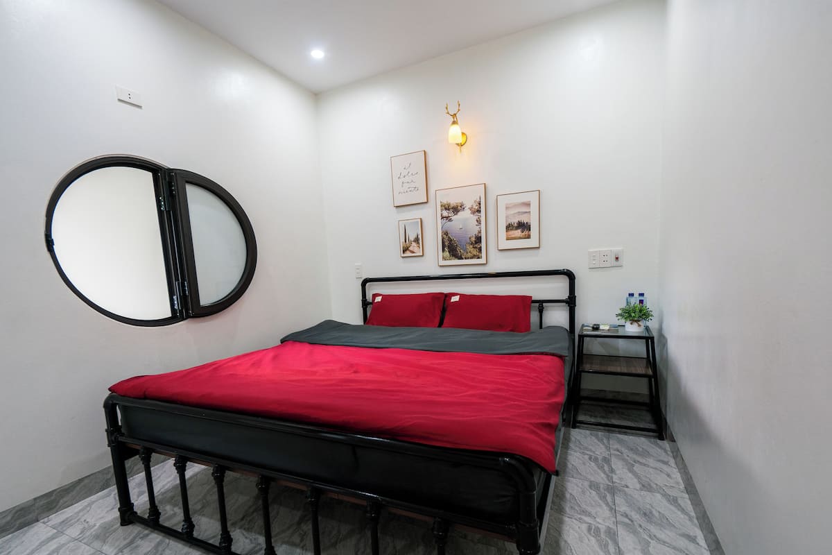 A bedroom with a bed, a round mirror, a bedside table with a plant, and framed pictures on the wall.
