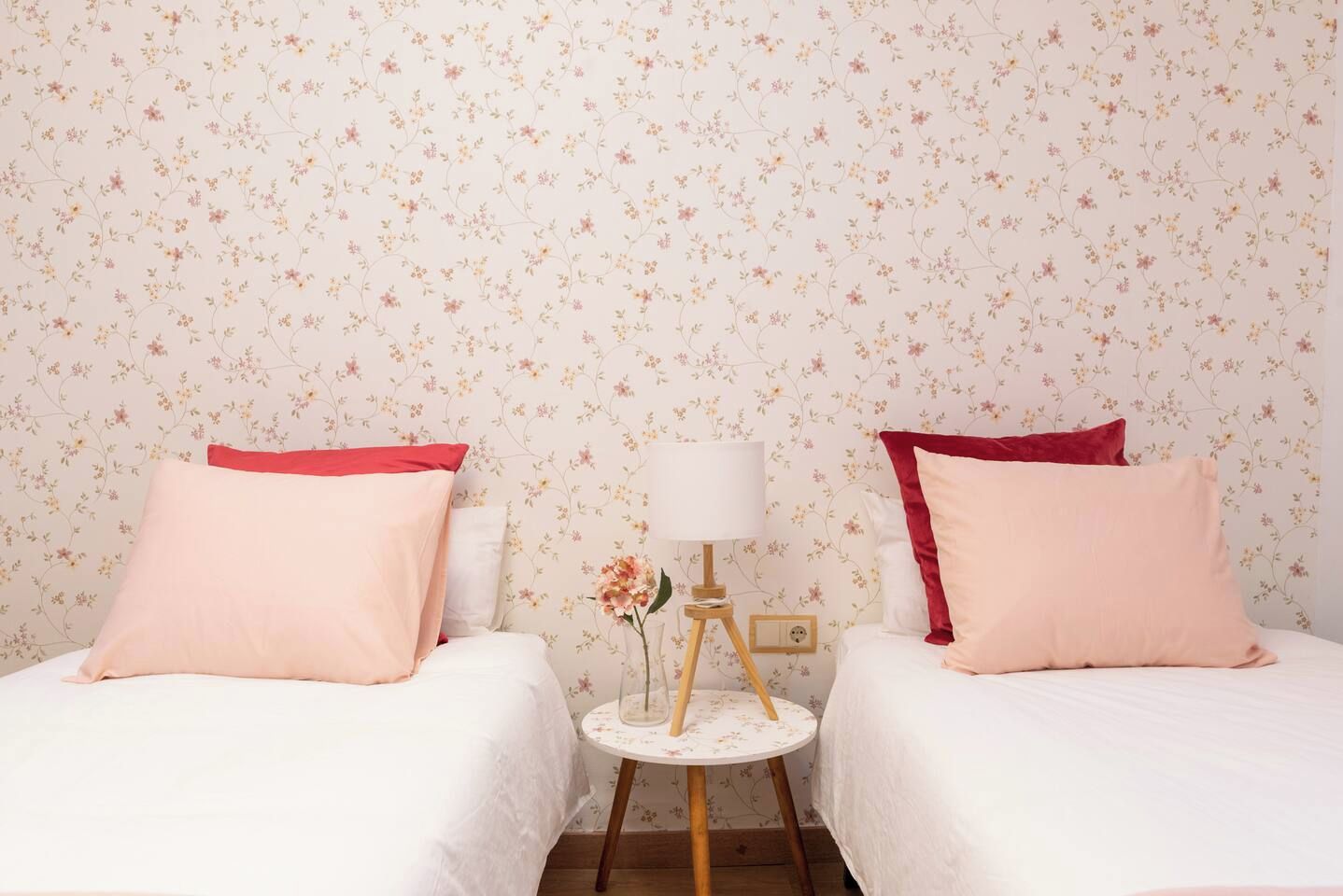 A room with two beds, each with a red accent pillow, a small round table with a lamp, and floral wallpaper.