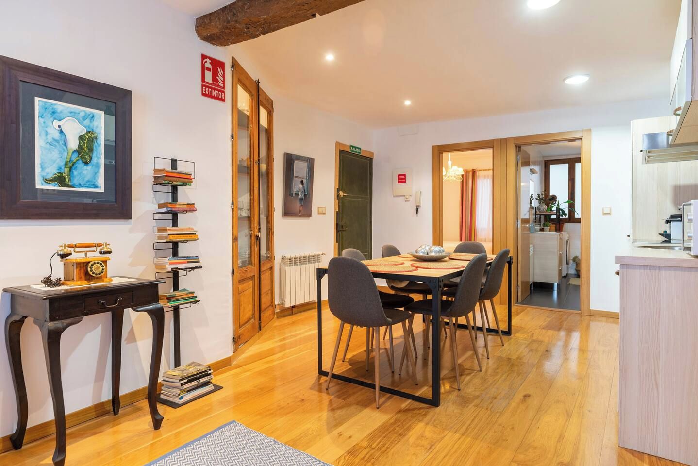 A dining area with a wooden table and chairs, a bookshelf with books, and a framed artwork on the wall.