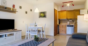 A living room with a TV, a dining table with chairs, a sofa, and a kitchen area with wooden cabinets and a dishwasher.
