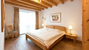 Ein Schlafzimmer mit einem hölzernen Bett, einem Sessel, einem Nachttisch mit Lampe, einem Heizkörper und einem Fenster mit Vorhängen.