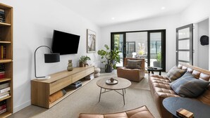 Een moderne woonkamer met een flatscreen televisie, een houten boekenkast, een leren bank, een bijzettafeltje en een grote glazen schuifdeur die leidt naar een buitenruimte.