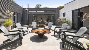 Een modern terras buiten met Adirondack-stoelen, een centraal vuurkorf en een eethoek met een tafel en stoelen.