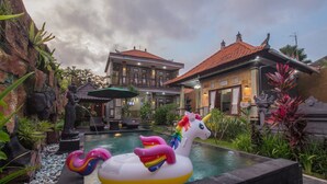 A pool area with a unicorn float, surrounded by greenery and a building with a tiled roof. - Win House Ubud by Supala (Ubud)