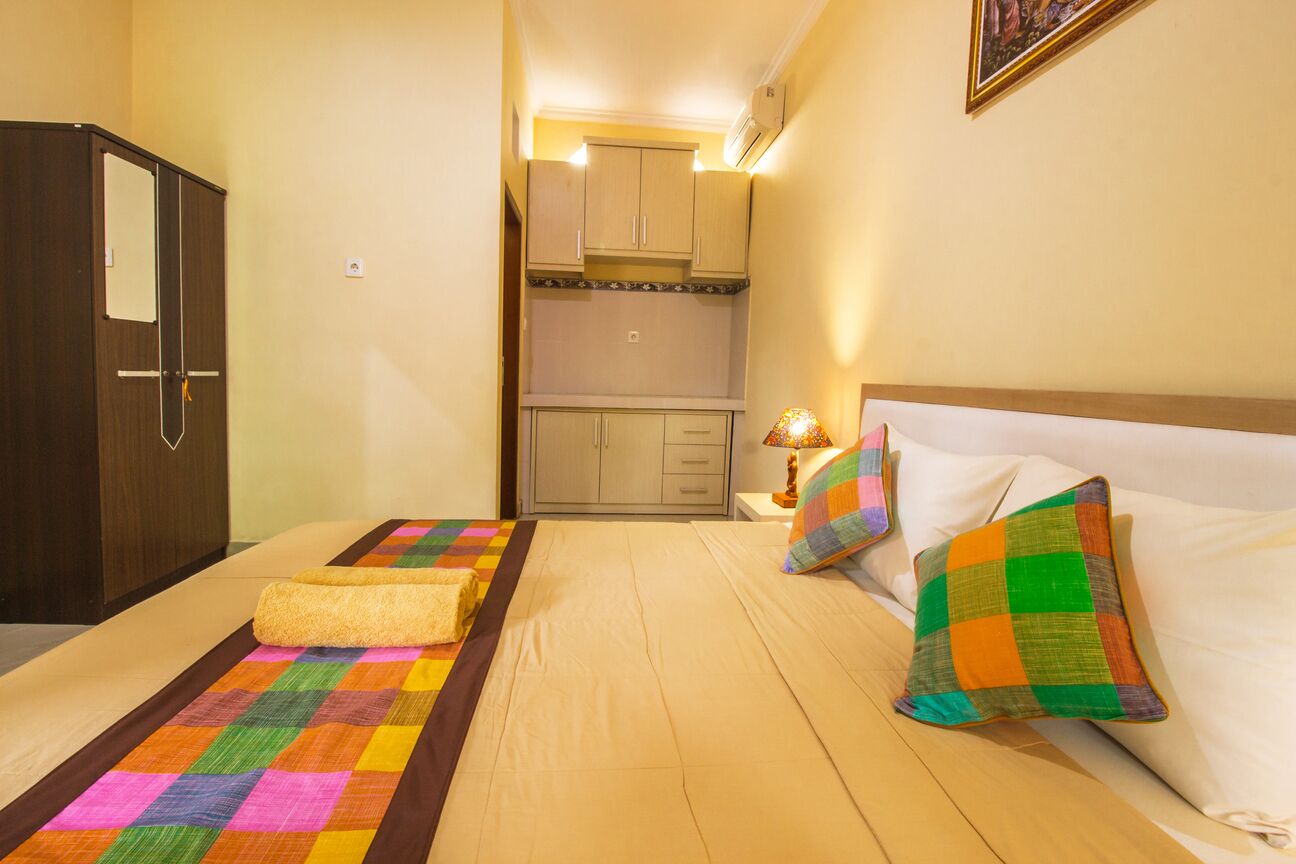 A hotel room with a bed, a colorful quilt, a pillow, a nightstand, a lamp, and a kitchenette in the background.