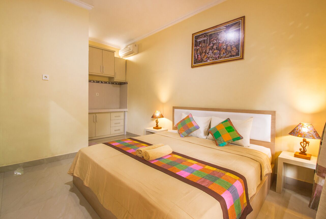 A hotel room with a bed, two bedside lamps, a nightstand, a painting on the wall, and a kitchen area visible through an open door.