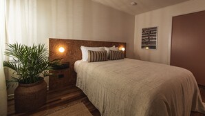 A bedroom with a large bed, a nightstand with a lamp, a potted plant, and a wall-mounted artwork.