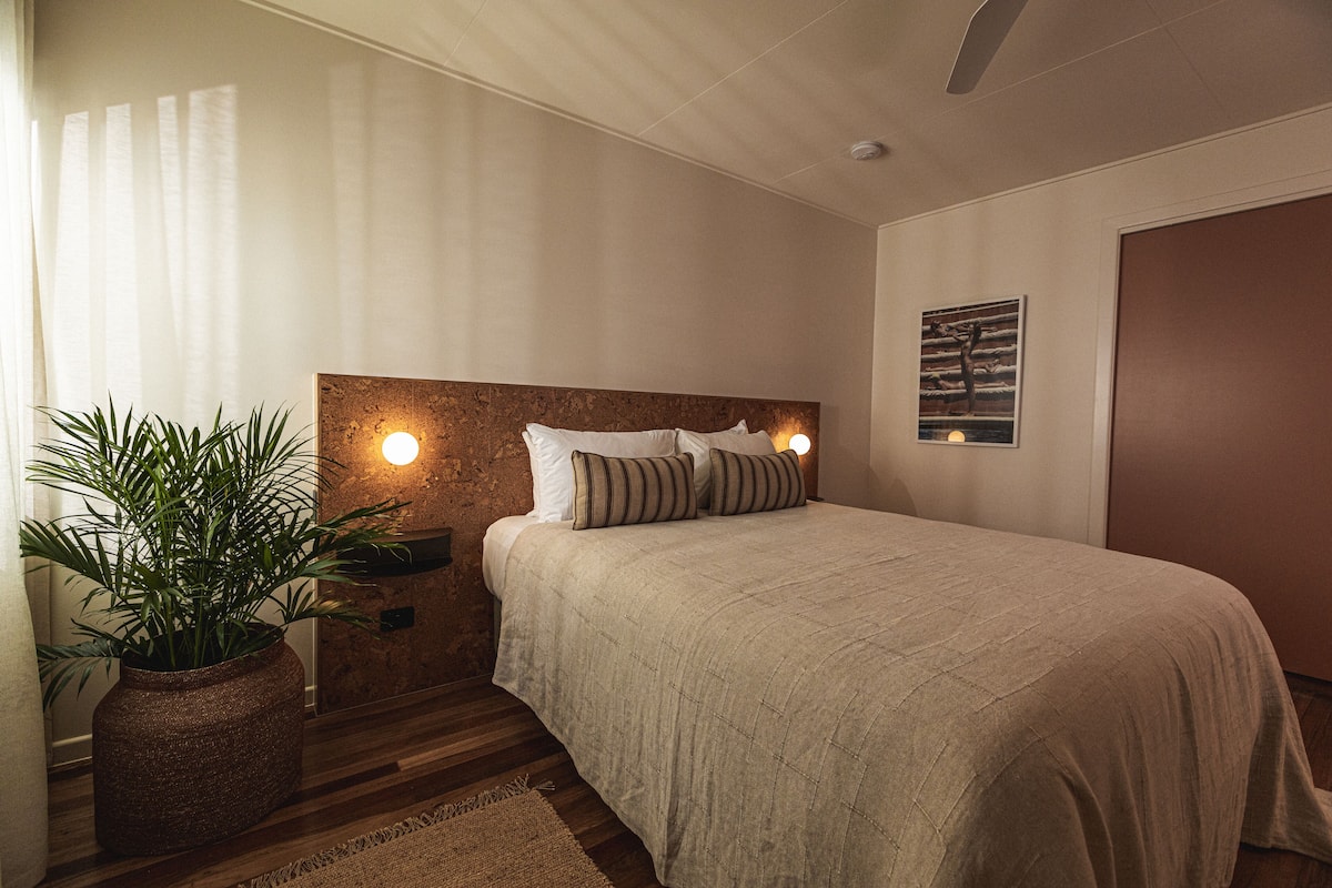 Een slaapkamer met een groot bed, een nachtkastje met een lamp, een potplant en een wanddecoratie.