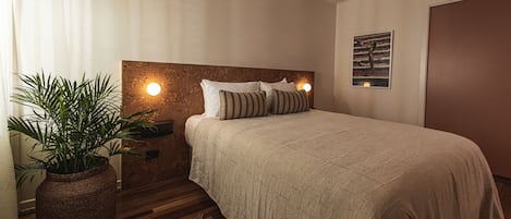 A bedroom with a large bed, a nightstand with a lamp, a potted plant, and a wall-mounted artwork.