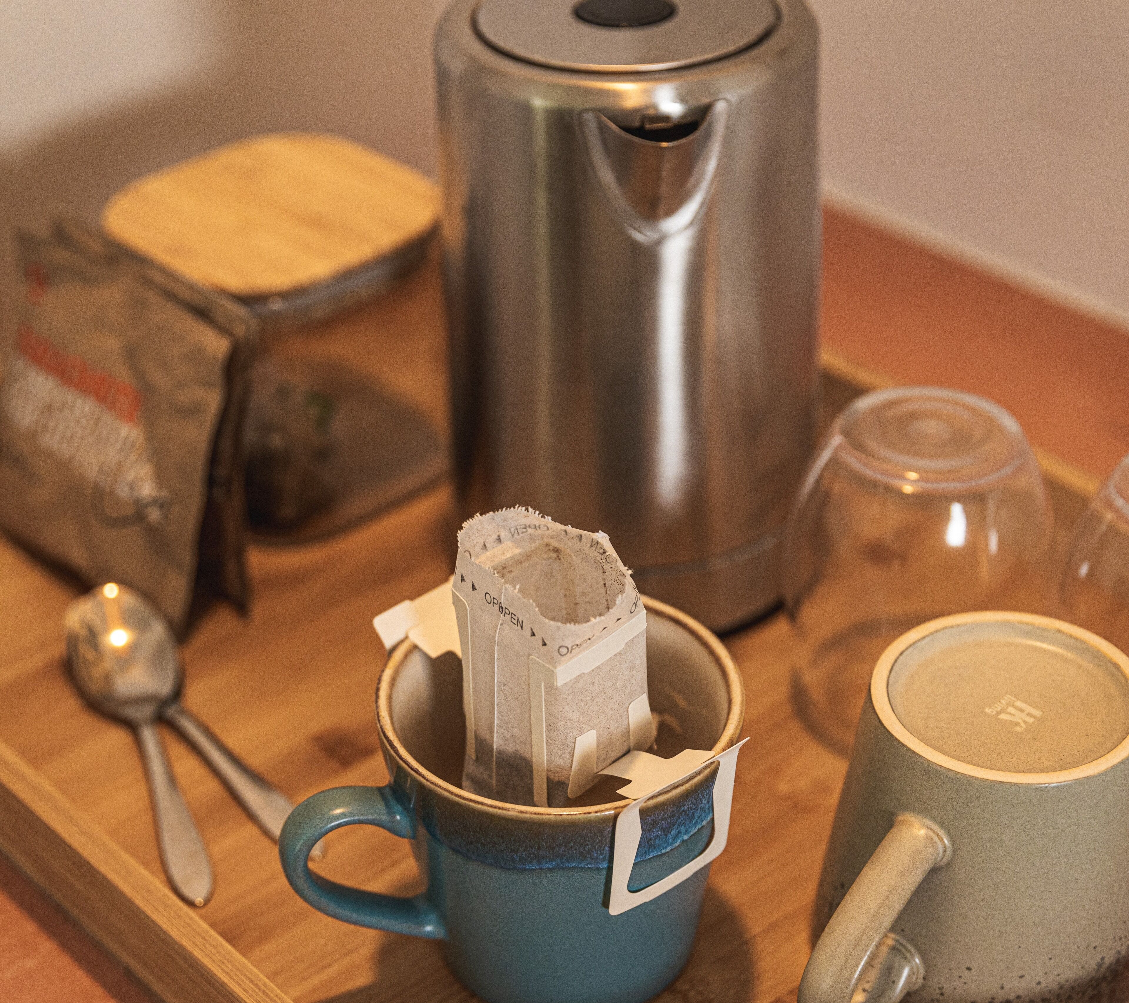 A blue mug with a tea bag, a stainless steel kettle, a glass cup, and a spoon on a wooden tray.