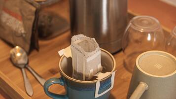 Una taza azul con una bolsita de té, una tetera de acero inoxidable, una taza de vidrio y una cuchara sobre una bandeja de madera.