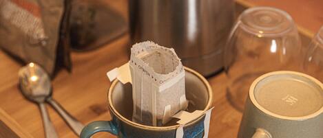 A blue mug with a tea bag, a stainless steel kettle, a glass cup, and a spoon on a wooden tray.
