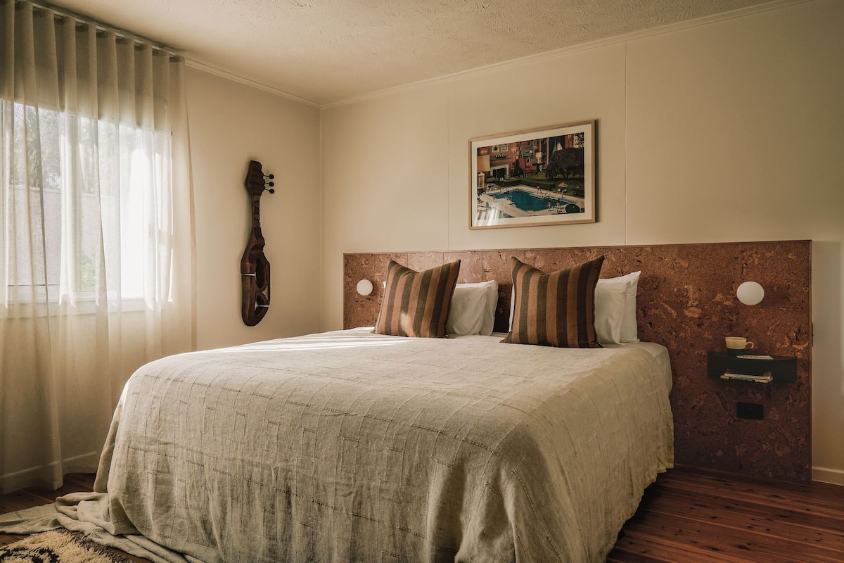 Een netjes opgemaakt bed met een hoofdeinde, een gitaar die tegen de muur staat en een ingelijste foto erboven.