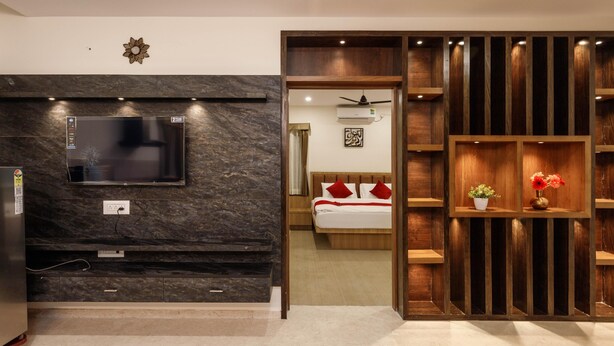 Een moderne hotelkamer met een flatscreen televisie bevestigd aan een donkere marmeren muur, een houten bed met rode kussens, en een houten wandplank met bloemen.