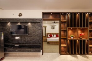 A modern hotel room with a flat-screen TV mounted on a dark marble wall, a wooden bed with red pillows, and a wooden shelving unit with flowers. - The Signature Serviced Apartment (Bengaluru)