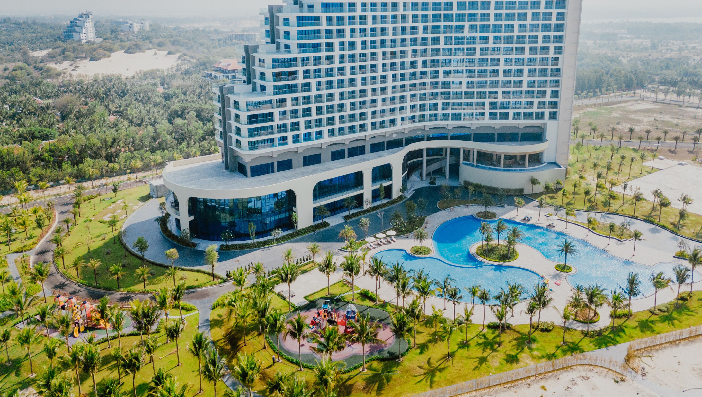 A modern hotel with a large pool area, surrounded by palm trees and greenery.