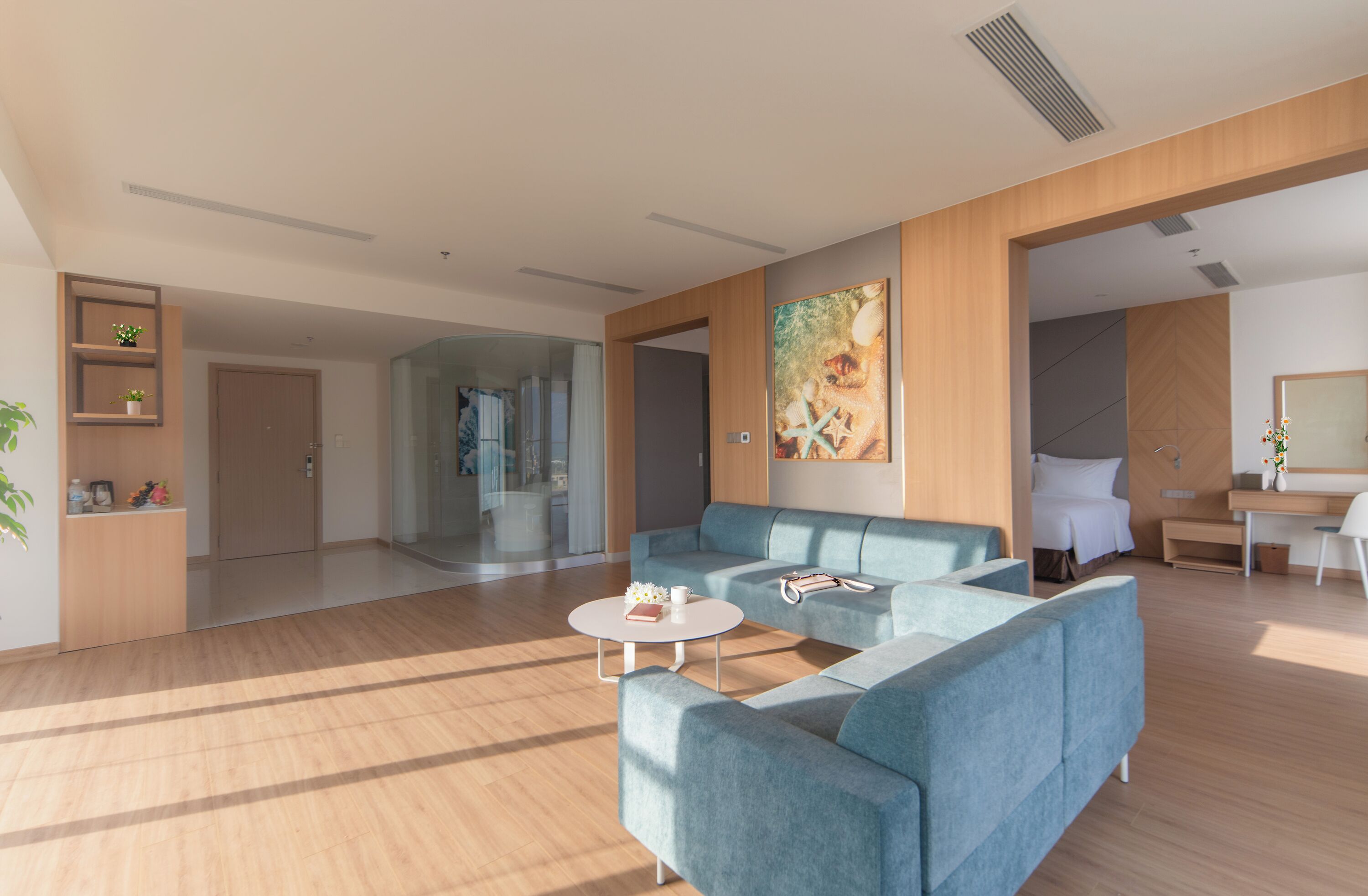 Uma sala de estar ampla com um sofá azul grande, uma mesa de centro e um quadro na parede. Há um armário de madeira com plantas e uma porta que dá para outro cômodo.