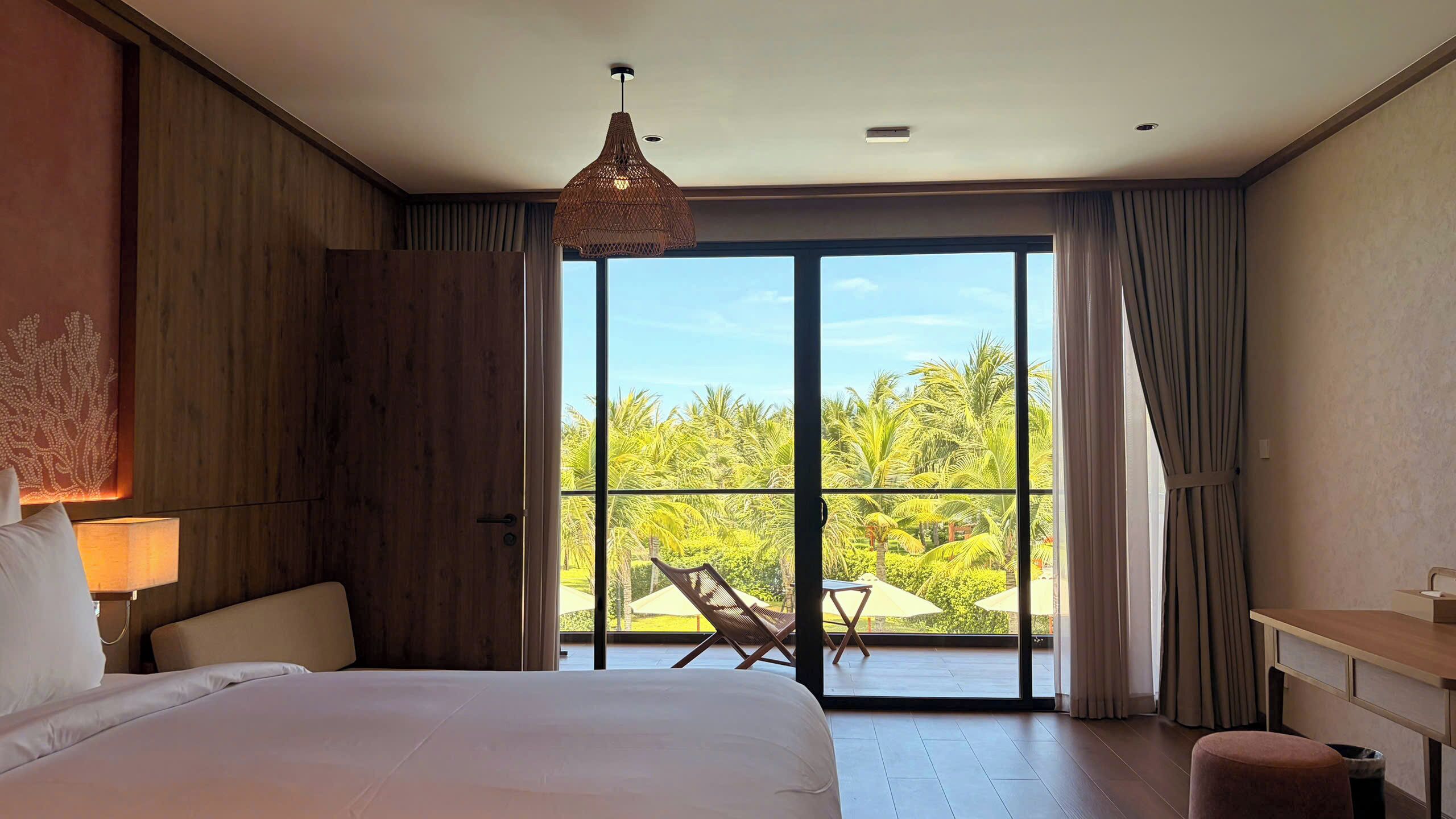 A hotel room with a large bed, a desk, a chair, and a view of palm trees through the glass door.