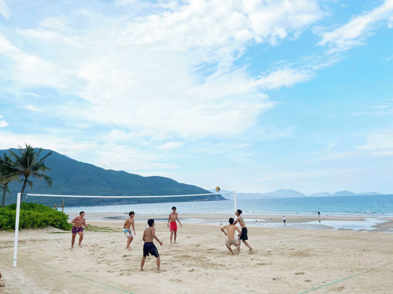 En strandvolleybollmatch på en sandplan med ett nät, palmer och berg i bakgrunden.