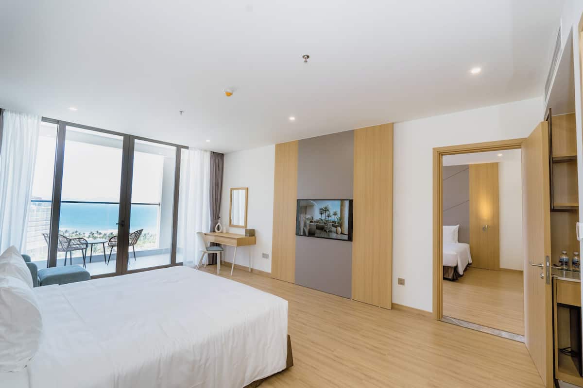 A modern hotel room with a large bed, a desk, and a TV. There is a balcony with a view of the sea.