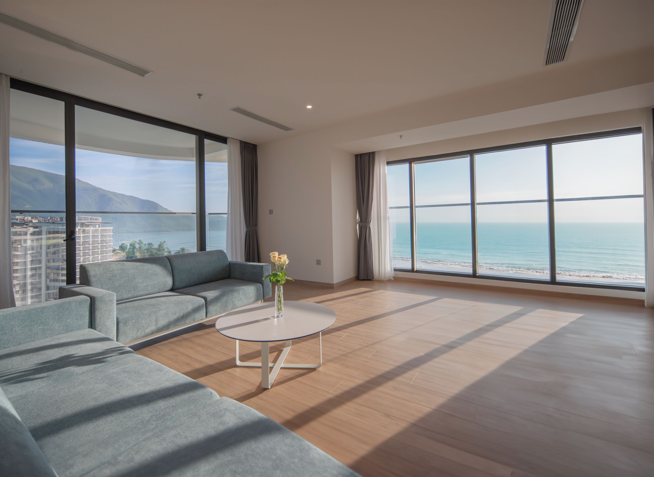 A modern living room with a grey sofa, a white coffee table, and a view of the sea and mountains.