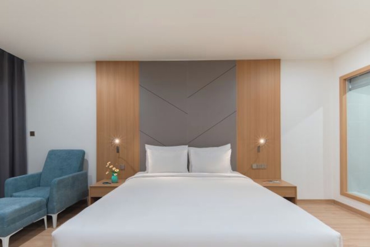 A modern hotel room with a large bed, a blue armchair, two bedside tables with lamps, and a large window.