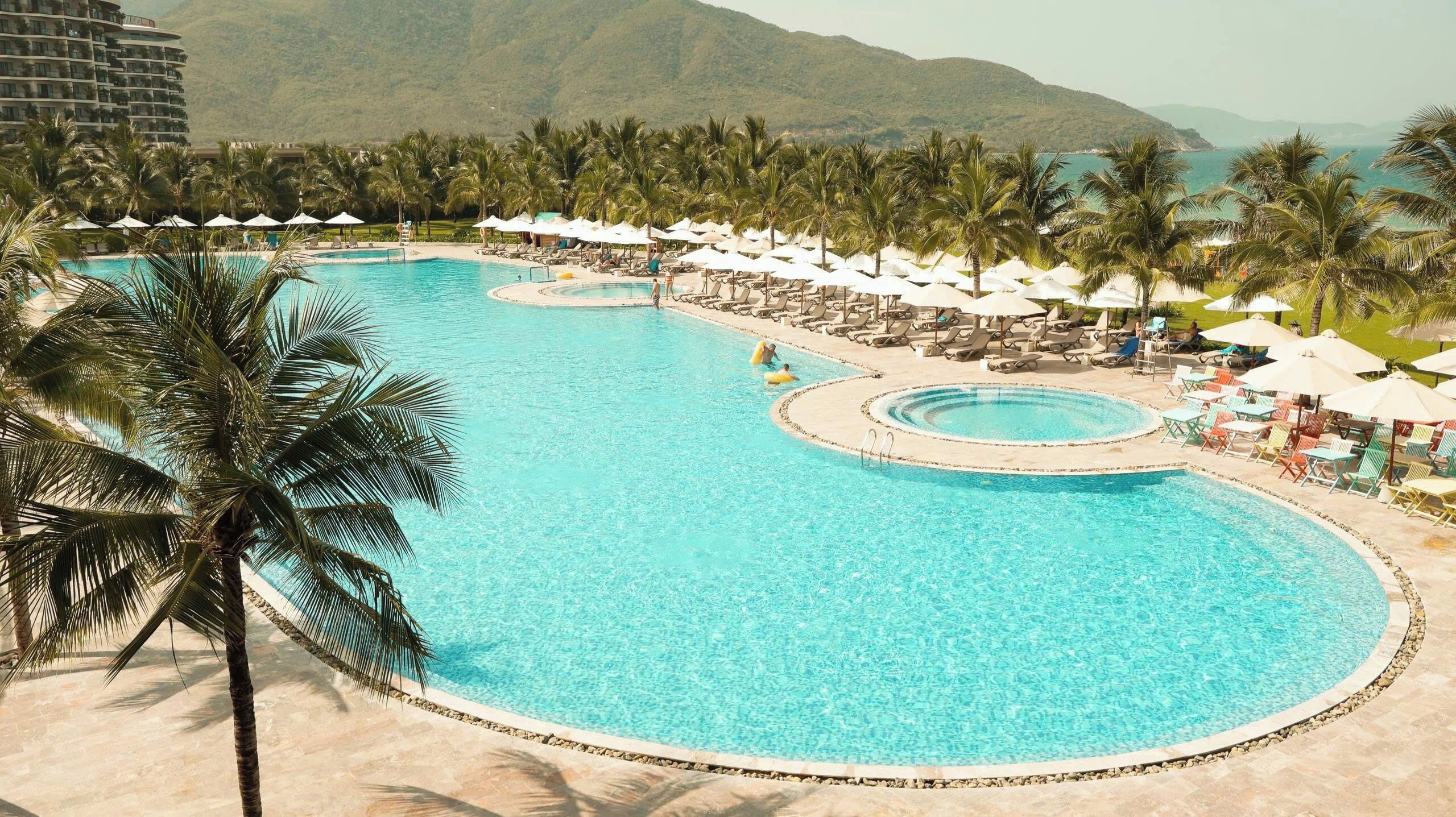 Una piscina amplia y cristalina con varias zonas de sol y sombrillas, rodeada de palmeras y montañas al fondo.
