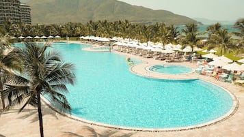 Una piscina amplia y cristalina con varias zonas de sol y sombrillas, rodeada de palmeras y montañas al fondo.
