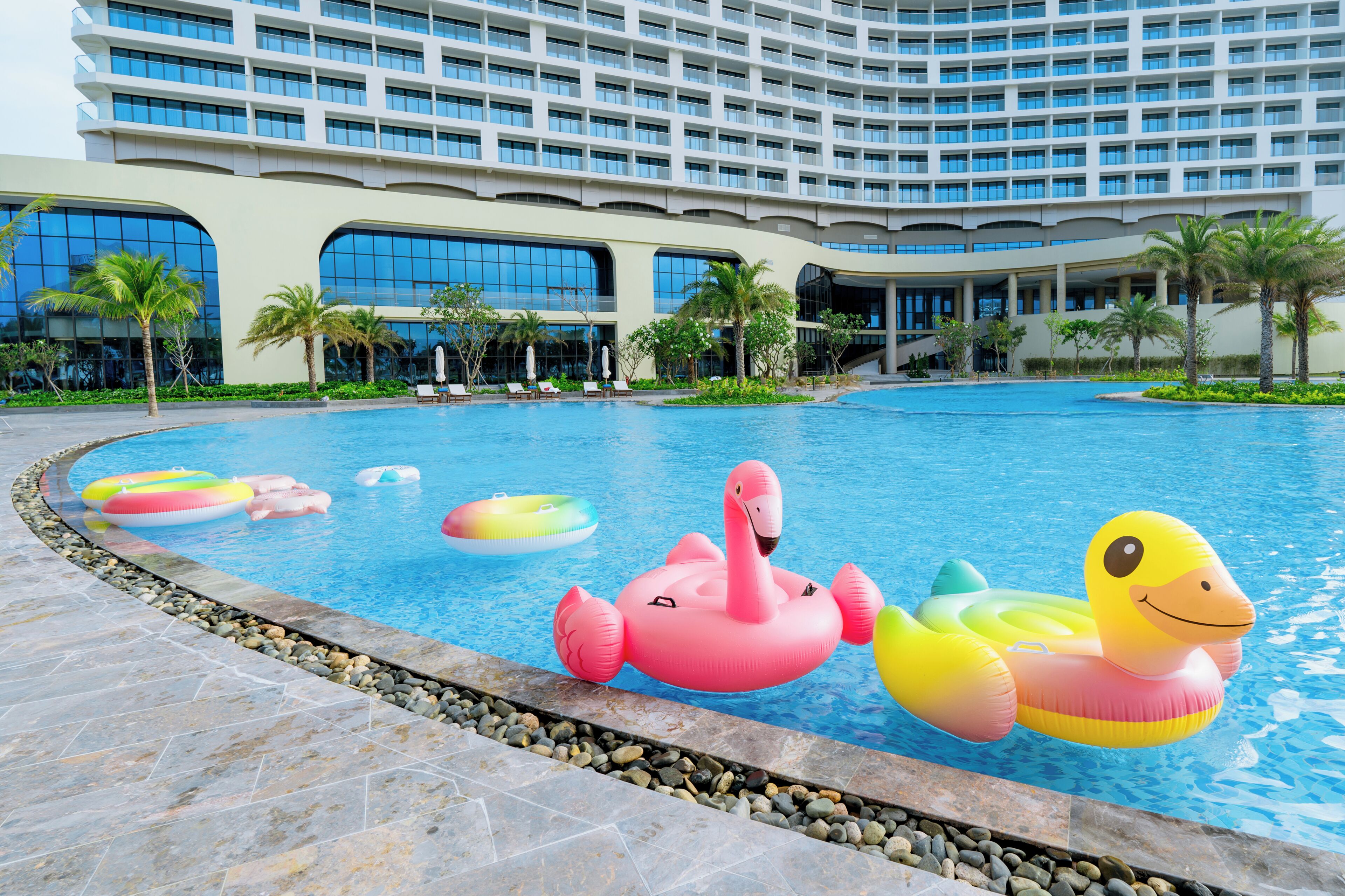 Une piscine d’hôtel avec de nombreux jeux gonflables, des palmiers et un grand bâtiment en arrière-plan.