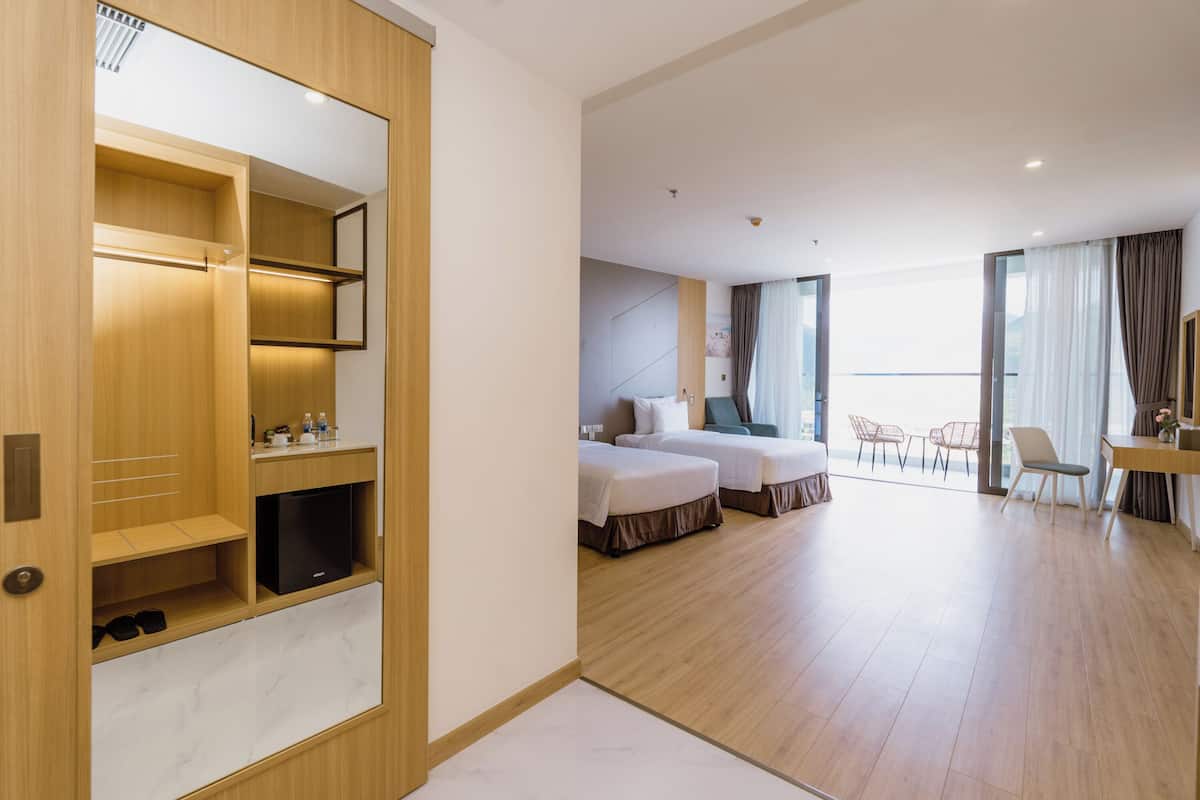A hotel room with a wooden wardrobe, two beds, a desk, and a view of the city.