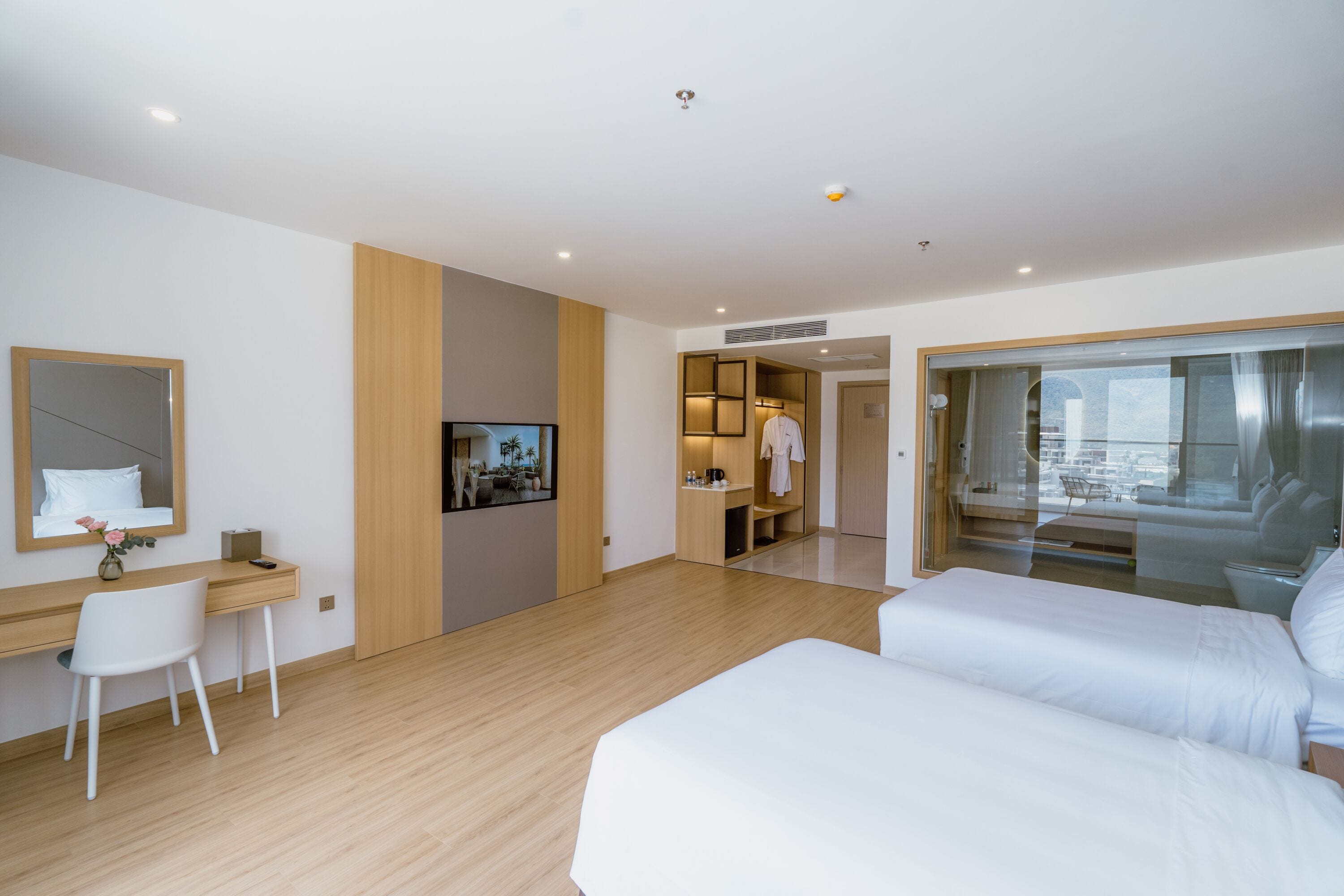 Une chambre d’hôtel moderne dotée d’un bureau en bois, d’une télévision, d’un lit et d’une porte vitrée donnant sur un balcon avec vue sur la ville.