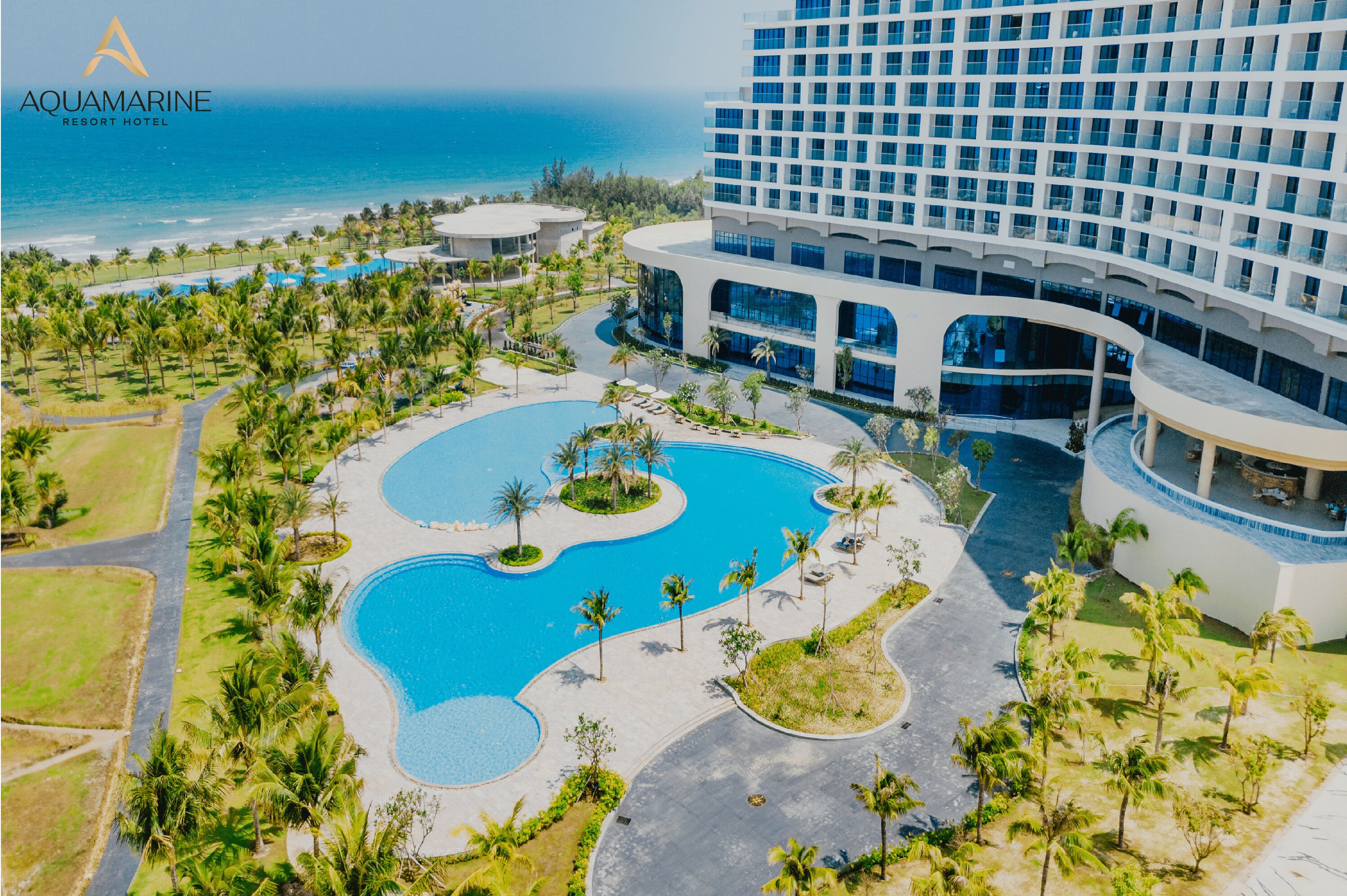 An aerial view of Aquamarine Resort Hotel with a large pool area, palm trees, and a view of the ocean.