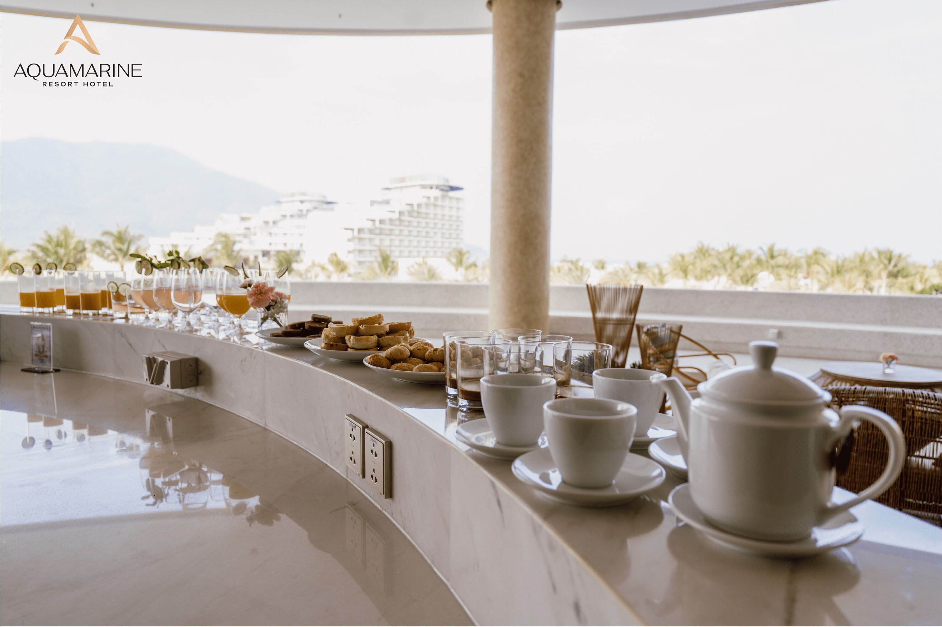 A breakfast buffet setup with various food items and beverages, a white teapot, and a view of buildings and palm trees in the background.